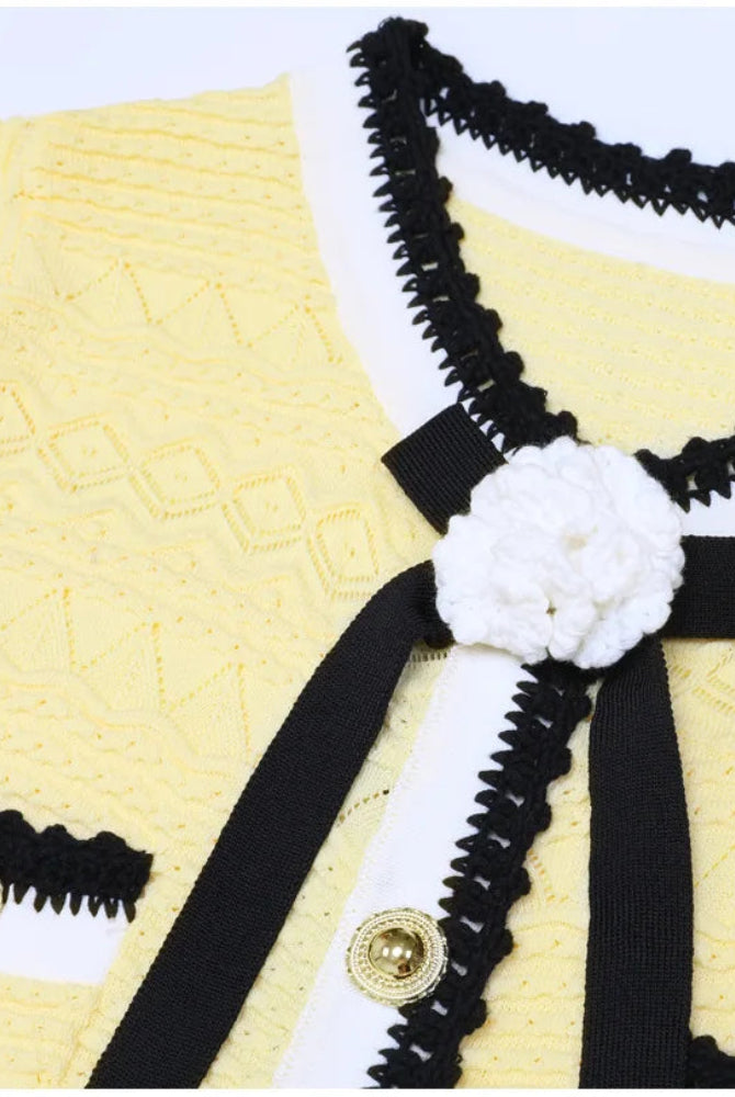 A close-up of a yellow knit cardigan featuring black and white trim, a black ribbon bow, and a white crocheted flower accent on the chest.