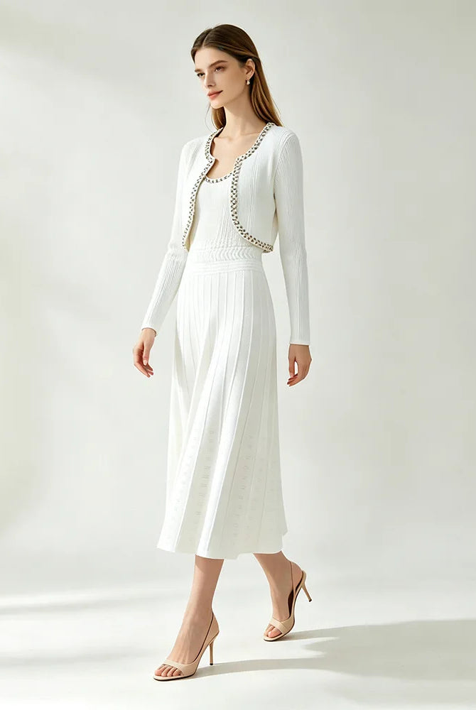 A woman models a white long-sleeve knit cardigan and pleated midi dress set with a decorative beaded neckline trim, paired with nude high heels, against a bright, minimalist studio background.