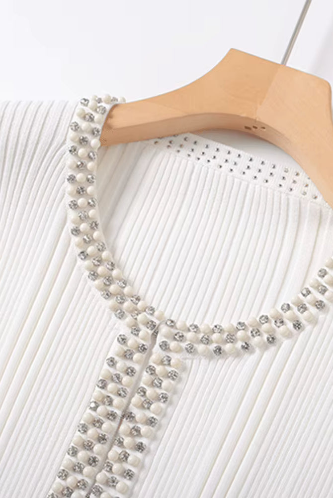 A close-up of a white ribbed knit top on a wooden hanger, featuring a decorative neckline and front trim embellished with white pearls and silver rhinestones.