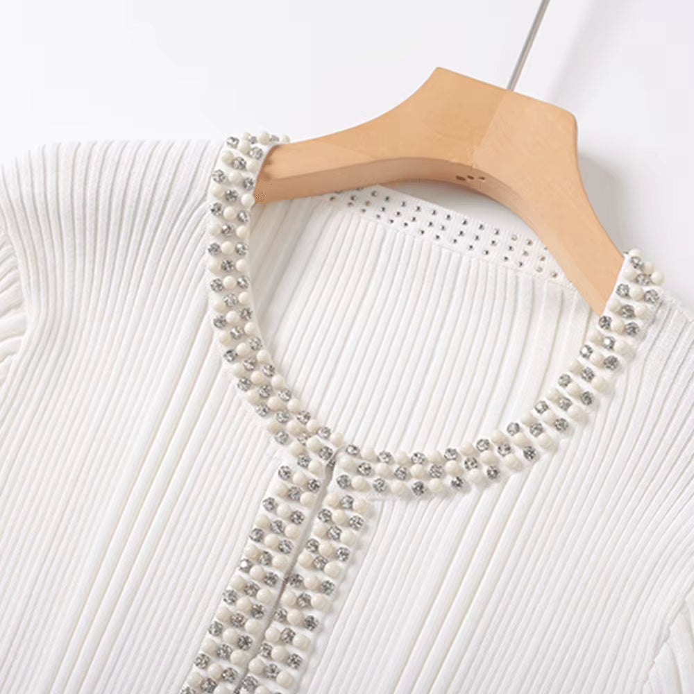 A close-up of a white ribbed knit top on a wooden hanger, featuring a decorative neckline and front trim embellished with white pearls and silver rhinestones.