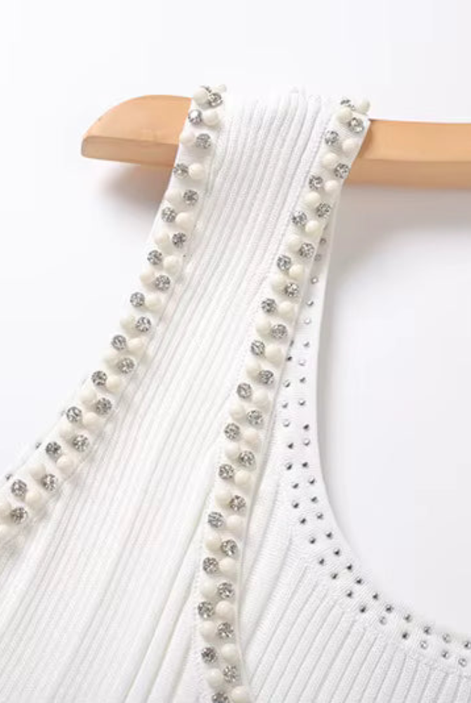 Close-up of a white ribbed dress with decorative straps featuring white pearls and silver rhinestones, hanging on a light wood hanger against a white background.