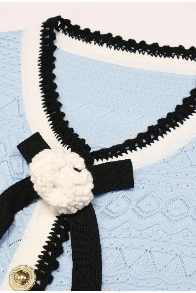 A close-up of a light blue textured knit cardigan featuring black and white trim, a black ribbon tie with a white crocheted flower accent, and gold-tone buttons.