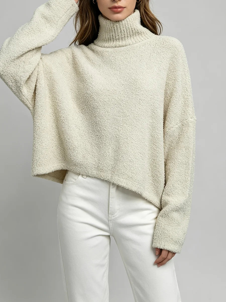 A woman wearing a cream-colored, textured boucle turtleneck sweater and white pants, shown from the chest down against a neutral gray background.