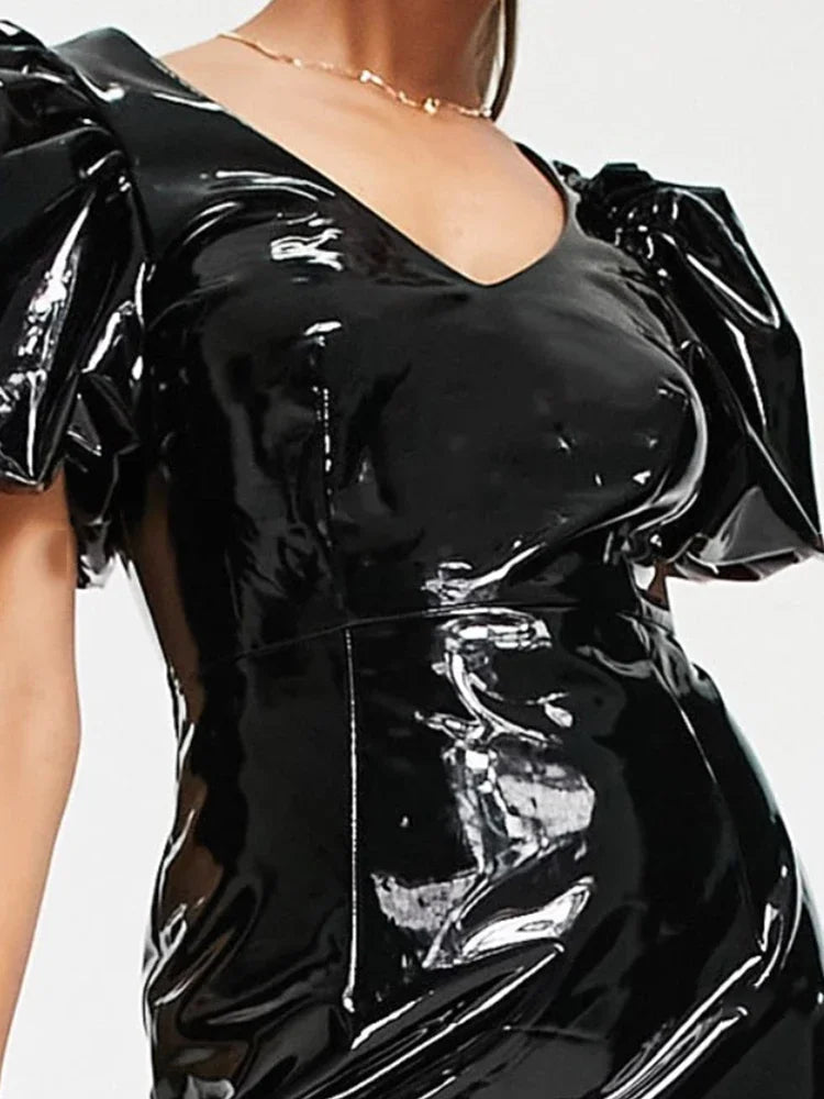 A close-up of a model wearing a shiny black latex or vinyl dress with dramatic puffed sleeves and a deep V-neckline.