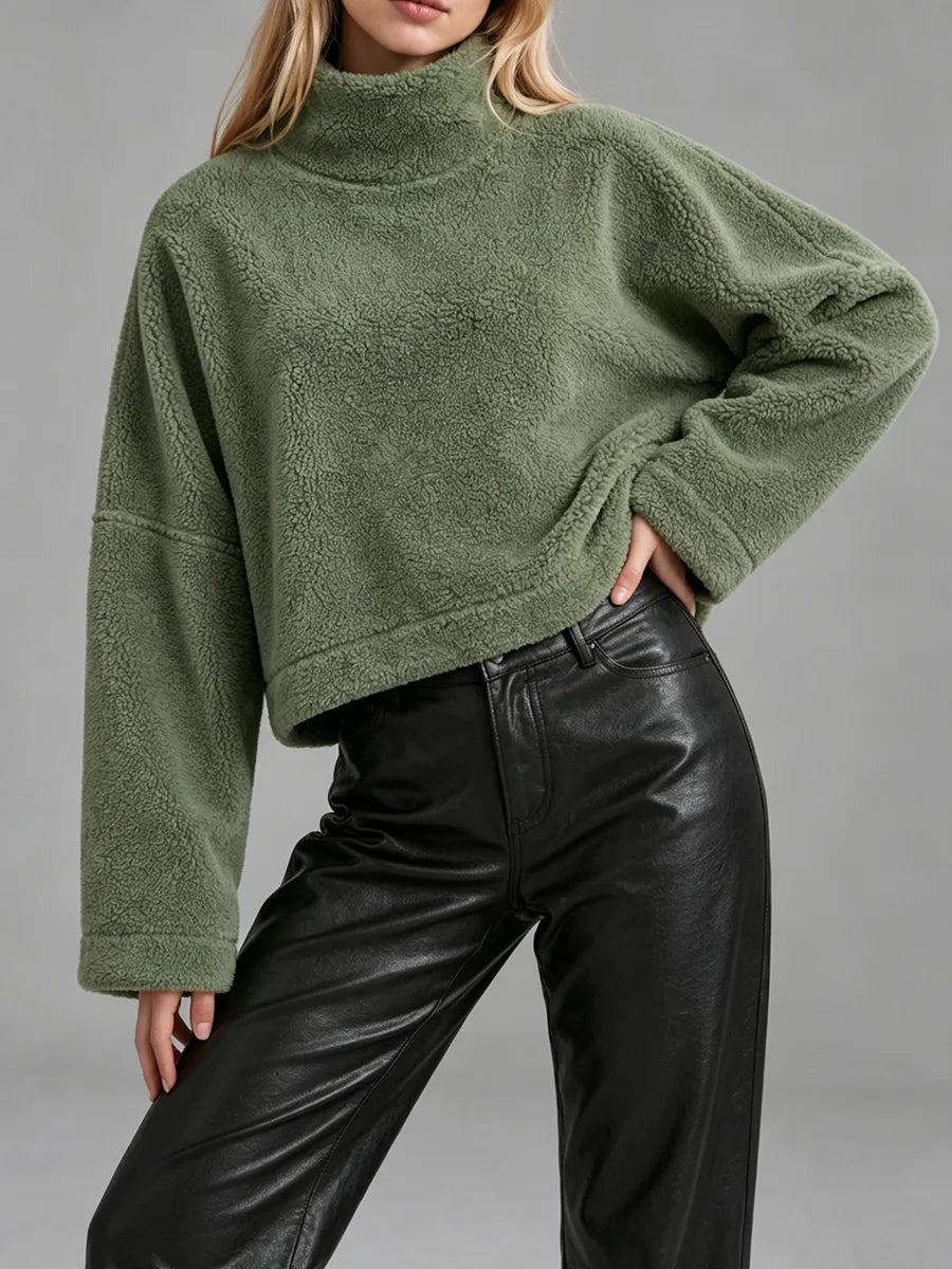 A woman wearing an oversized, cropped green sherpa fleece mock-neck sweater paired with black leather pants, shown against a neutral gray studio background.