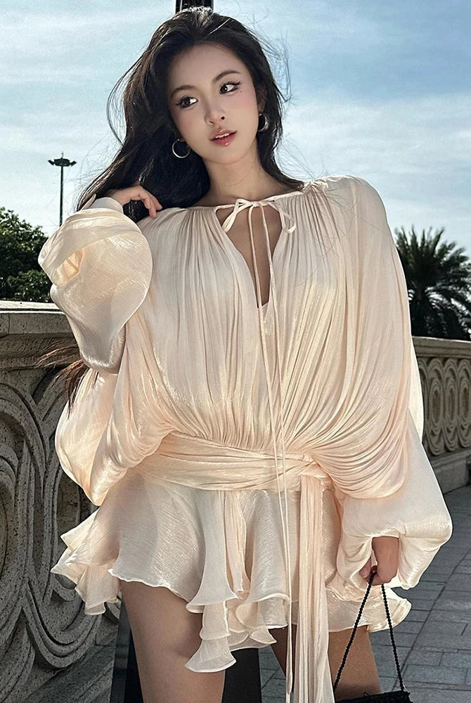 Woman wearing a cream ruched long sleeve mini dress with tie neckline and ruffle hem