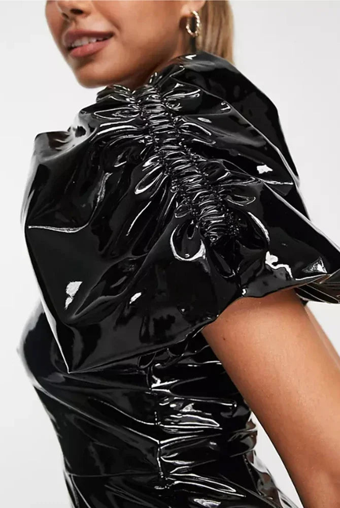 Close-up of a model wearing a glossy black vinyl or latex top with dramatic gathered puff sleeves and ruching, against a plain light background.