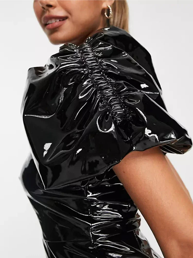 Close-up of a model wearing a glossy black vinyl or latex top with dramatic gathered puff sleeves and ruching, against a plain light background.