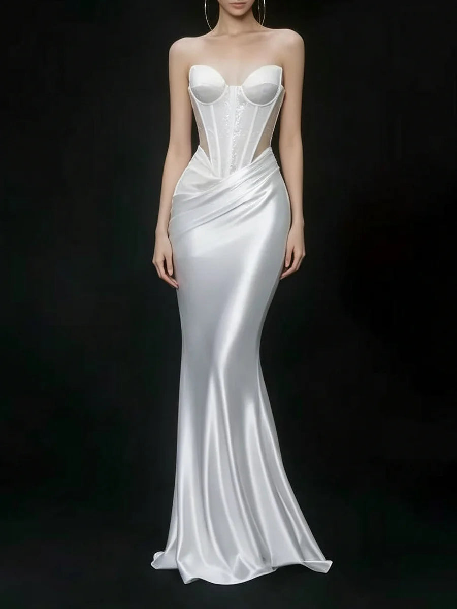 A strapless white satin gown with a structured corset-style bodice, sheer side panels, and a fitted mermaid silhouette, shown on a model against a black studio background.