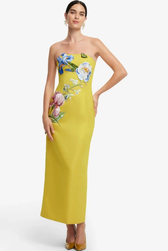 A woman modeling a strapless yellow maxi dress with colorful floral embroidery on the bodice and side, paired with gold heels against a light gray studio background.