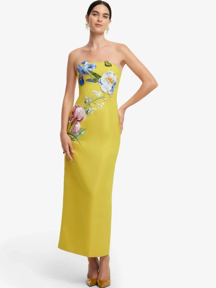 A woman modeling a strapless yellow maxi dress with colorful floral embroidery on the bodice and side, paired with gold heels against a light gray studio background.