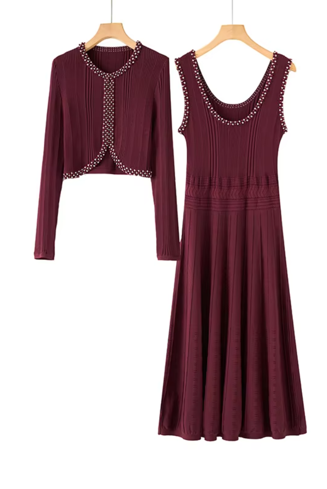 A two-piece women's outfit set in a deep burgundy color, featuring a long-sleeve cropped cardigan with decorative silver chain trim and a matching sleeveless midi dress with a scoop neckline and embellished shoulder straps, shown hanging on wooden hangers against a plain white background.