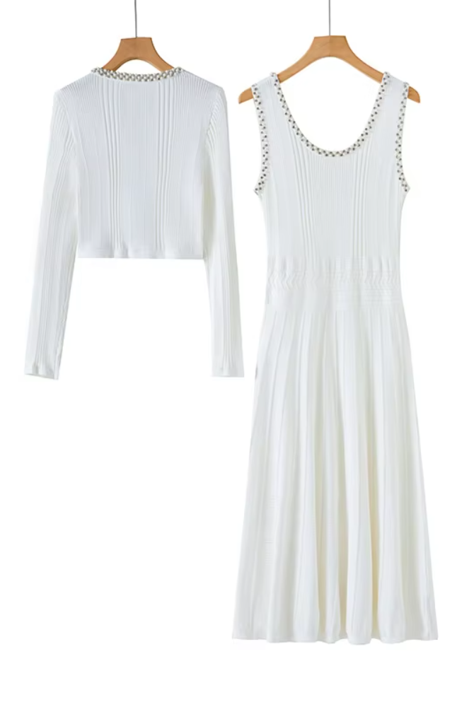 A white sleeveless midi dress with a pleated bodice and shoulder chain detailing, shown alongside a matching white long-sleeve cropped knit top on wooden hangers against a white background.