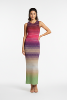 A woman modeling a sleeveless knit maxi dress with a horizontal ombre pattern transitioning from red and pink at the top, through purple and beige, to green at the bottom, against a plain white studio background.