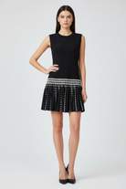 A woman modeling a sleeveless black mini dress with a pleated skirt and a horizontal black-and-white patterned band at the waist, photographed against a light gray studio background.
