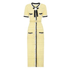 A pale yellow tweed midi dress with black and white trim, featuring a short-sleeve design, a black bow at the collar, a matching waist belt, and decorative buttons down the front.