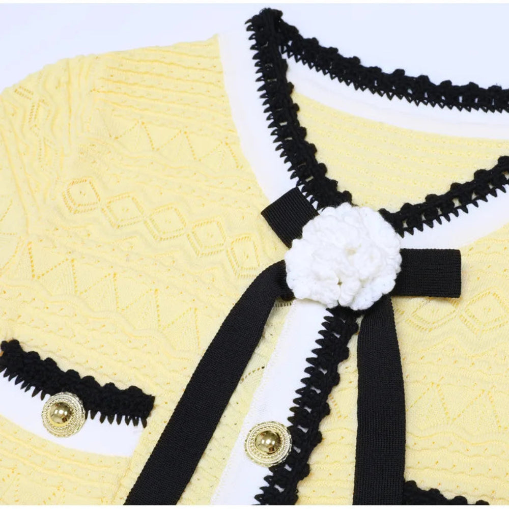 A close-up of a yellow knit cardigan featuring black and white trim, a black ribbon bow, and a white crocheted flower accent on the chest.