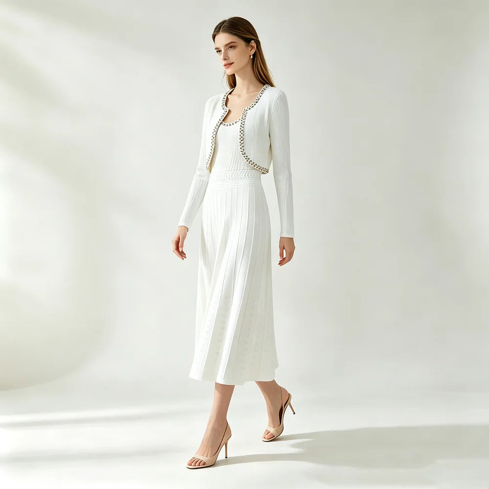 A woman models a white long-sleeve knit cardigan and pleated midi dress set with a decorative beaded neckline trim, paired with nude high heels, against a bright, minimalist studio background.