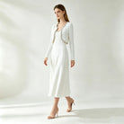 A woman models a white long-sleeve knit cardigan and pleated midi dress set with a decorative beaded neckline trim, paired with nude high heels, against a bright, minimalist studio background.