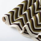 A close-up of a knitted fabric with a bold chevron pattern in olive green, dark brown, and cream, featuring ribbed knit borders.