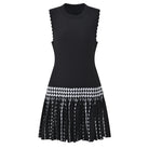A sleeveless black knit dress with scalloped armholes, a textured bodice, and a pleated skirt featuring a black and white harlequin diamond pattern band and fringe.