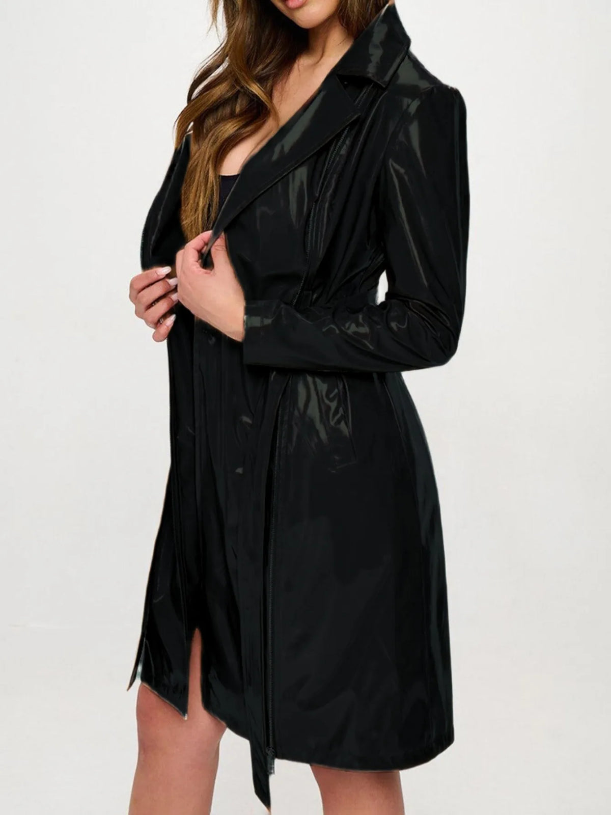 A woman wearing a shiny black longline trench coat over a black dress, shown from the neck down against a plain white studio background.