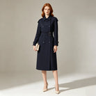 A woman stands in a studio wearing a dark navy knee-length trench coat with a belt.