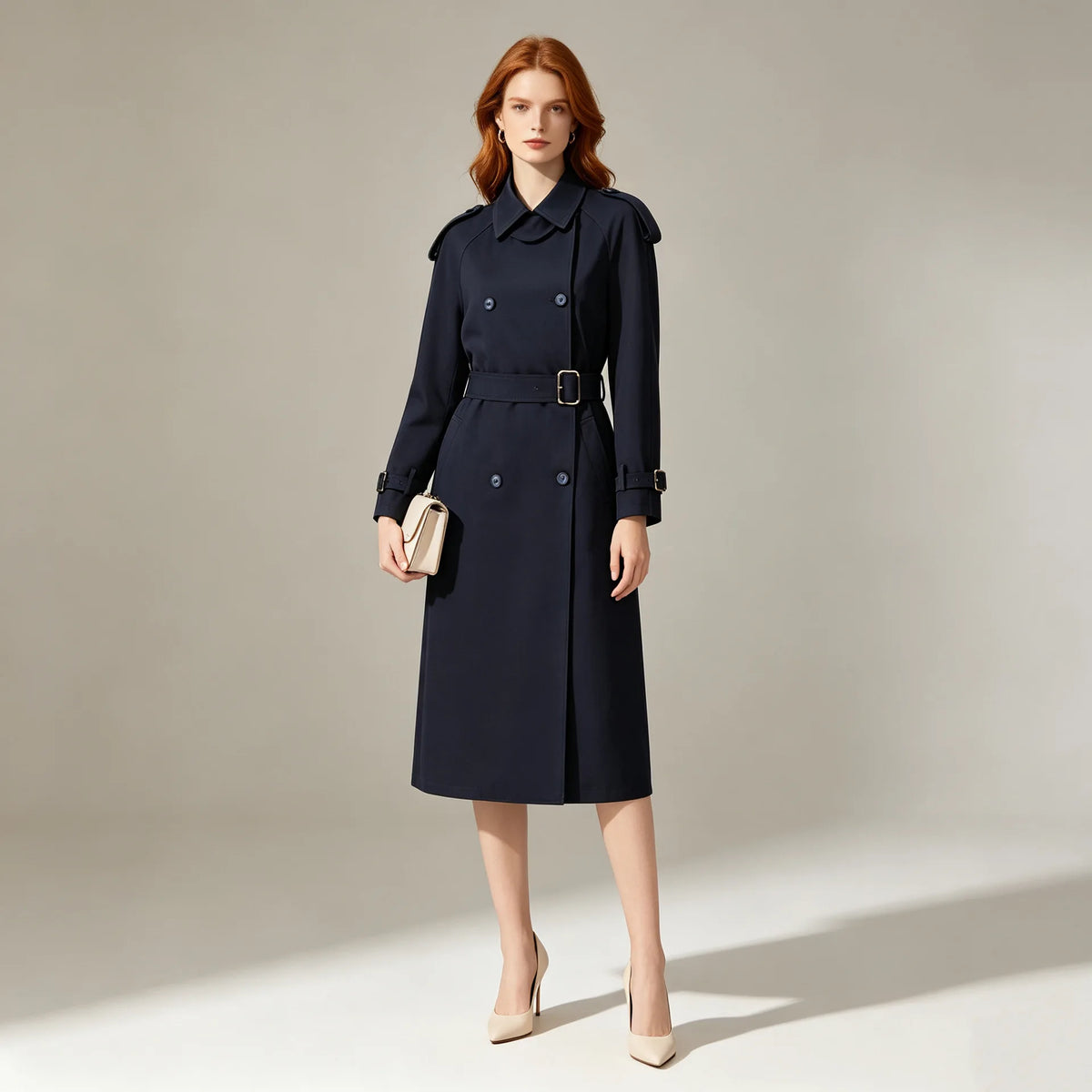 A woman stands in a studio wearing a dark navy knee-length trench coat with a belt.