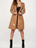 A woman modeling a glossy, shiny tan trench coat over a black dress and black patent leather ankle boots, standing against a plain white studio background.