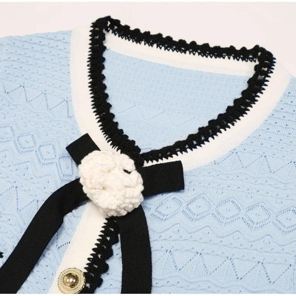 A close-up of a light blue textured knit cardigan featuring black and white trim, a black ribbon tie with a white crocheted flower accent, and gold-tone buttons.