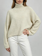 A woman wearing a cream-colored, textured boucle turtleneck sweater and white pants, shown from the neck down against a neutral gray background.
