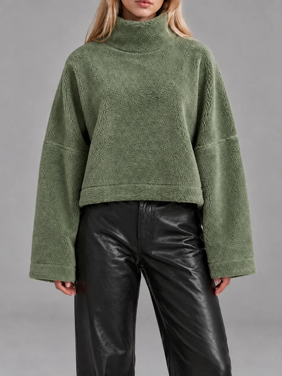 A model wearing a cropped, olive green textured fleece turtleneck sweater with wide long sleeves, paired with black leather pants, against a solid gray studio background.