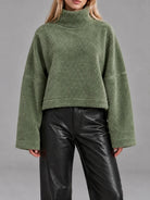 A model wearing a cropped, olive green textured fleece turtleneck sweater with wide long sleeves, paired with black leather pants, against a solid gray studio background.
