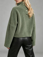 A woman wearing an olive green sherpa fleece mock neck pullover with a buttoned collar detail, paired with black leather pants, shown from a rear three-quarter angle against a neutral gray studio background.