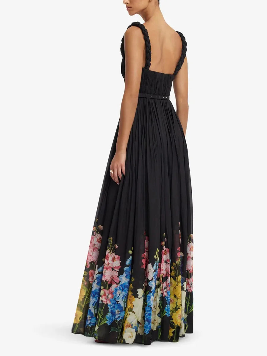 A woman modeling a long black maxi dress from the back, featuring a smocked bodice, ruffled straps, and a vibrant multicolor floral print along the hem against a light gray studio background.