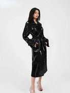 A woman modeling a glossy black long trench coat with a tie waist, photographed against a plain light gray studio background.