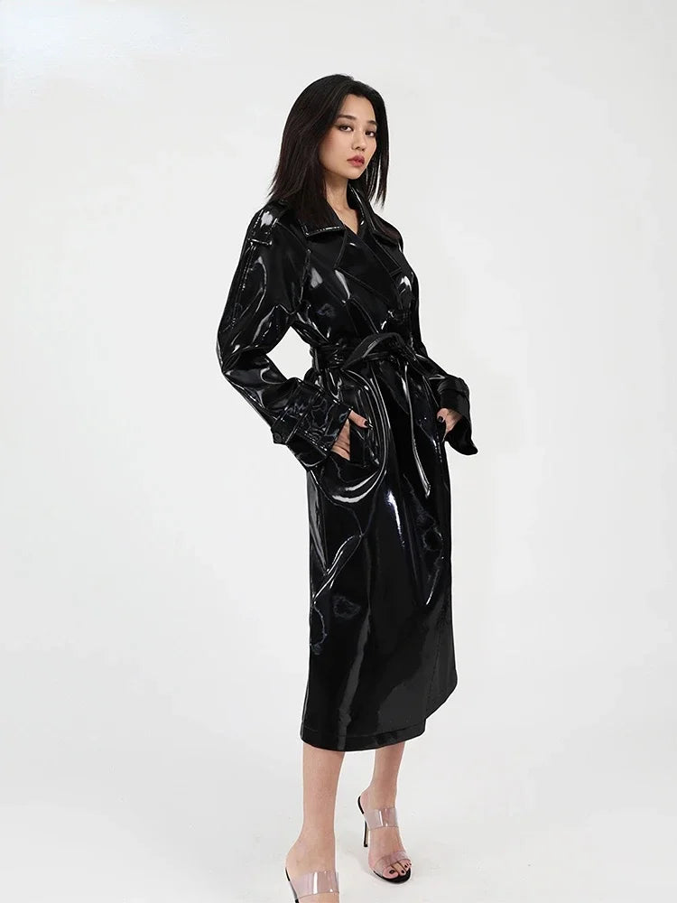 A woman modeling a glossy black long trench coat with a tie waist, photographed against a plain light gray studio background.