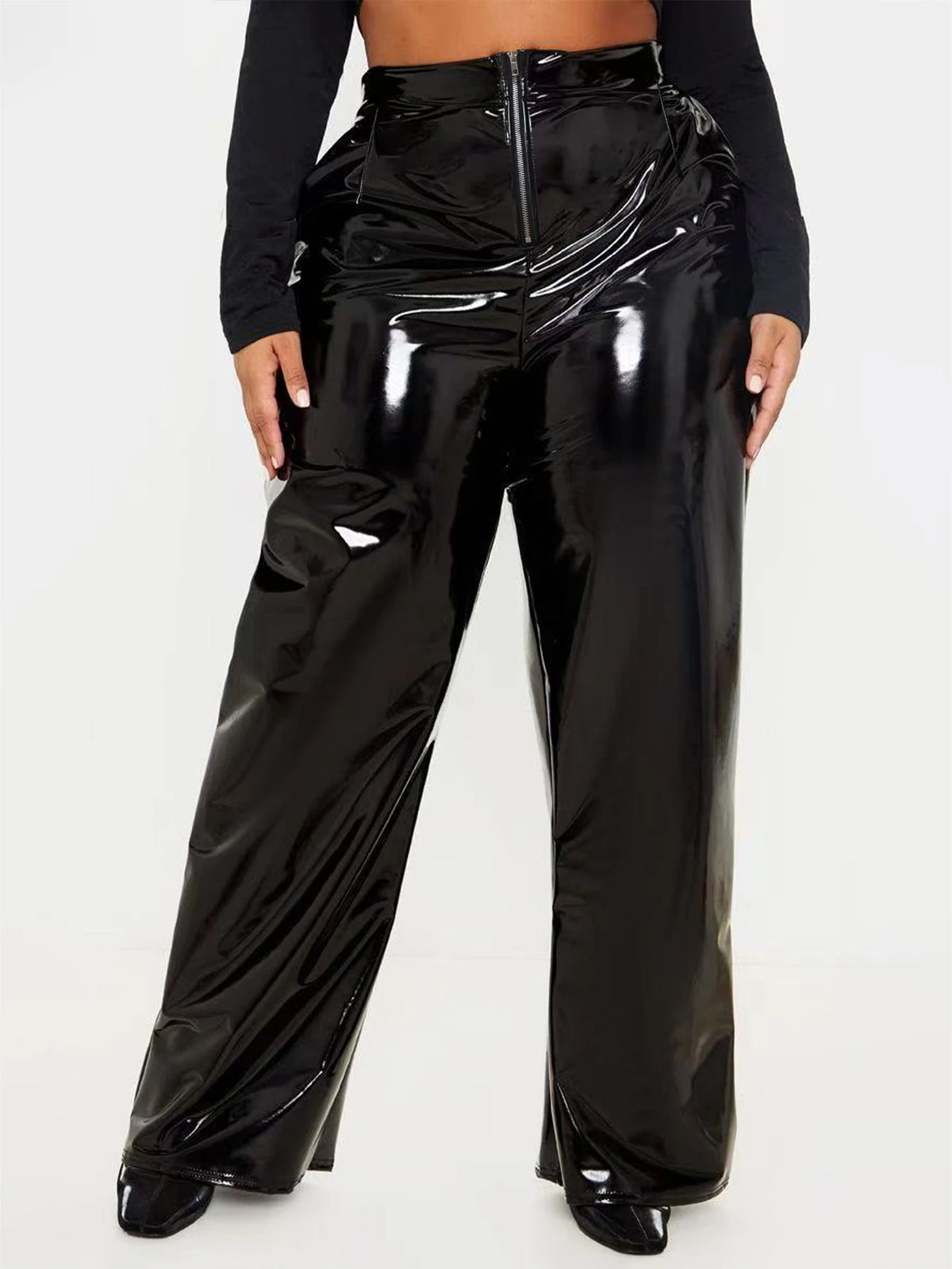 A model wearing high-waisted, wide-leg patent black pants with a front zipper, paired with a black long-sleeve top and black pointed shoes, against a plain white studio background.