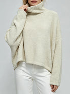A woman wearing a cream-colored, textured boucle turtleneck sweater and white pants, shown from the chin down against a light gray studio background.