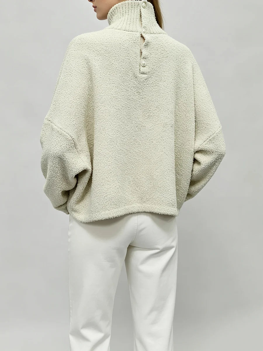 A person shown from the back wearing a cream-colored, textured boucle turtleneck sweater and white pants against a light gray studio background.