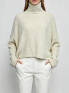 A woman wearing a cream-colored, fuzzy turtleneck sweater and white trousers, standing against a light gray studio background.