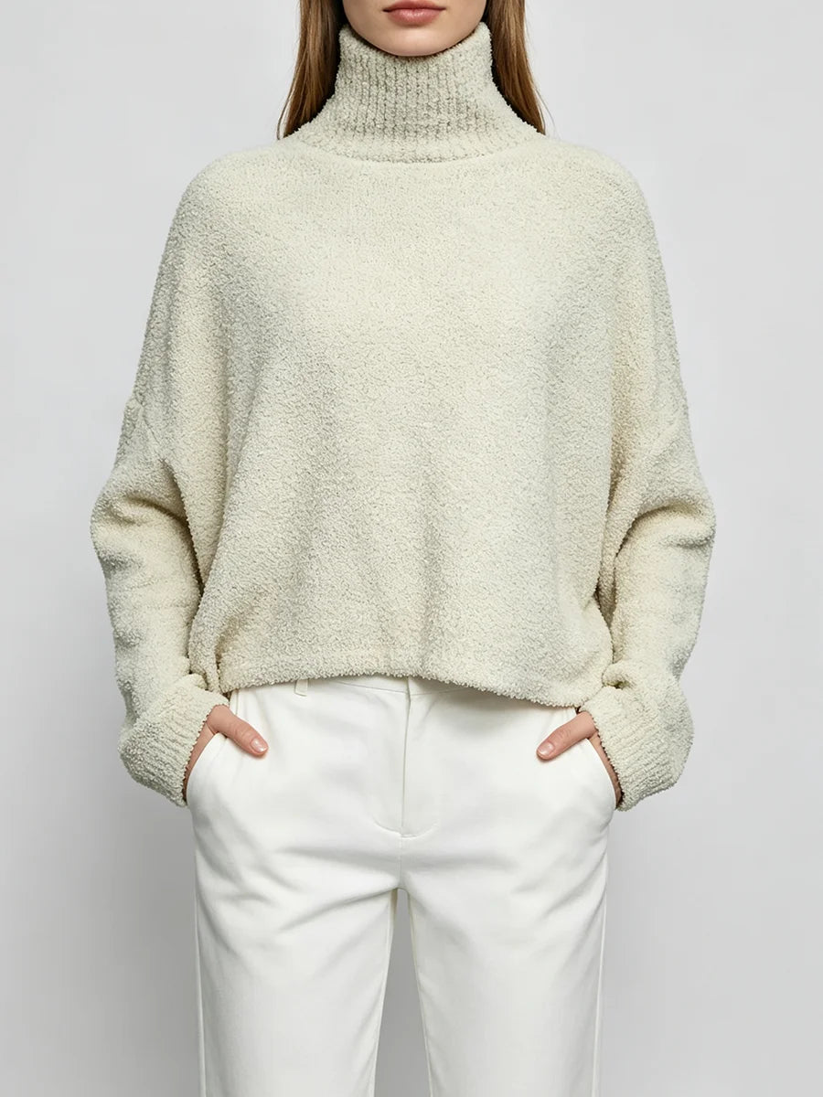 A woman wearing a cream-colored, fuzzy turtleneck sweater and white trousers, standing against a light gray studio background.