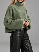A woman wearing a cropped, oversized green sherpa fleece turtleneck sweater and black faux-leather pants, photographed against a plain gray studio background.