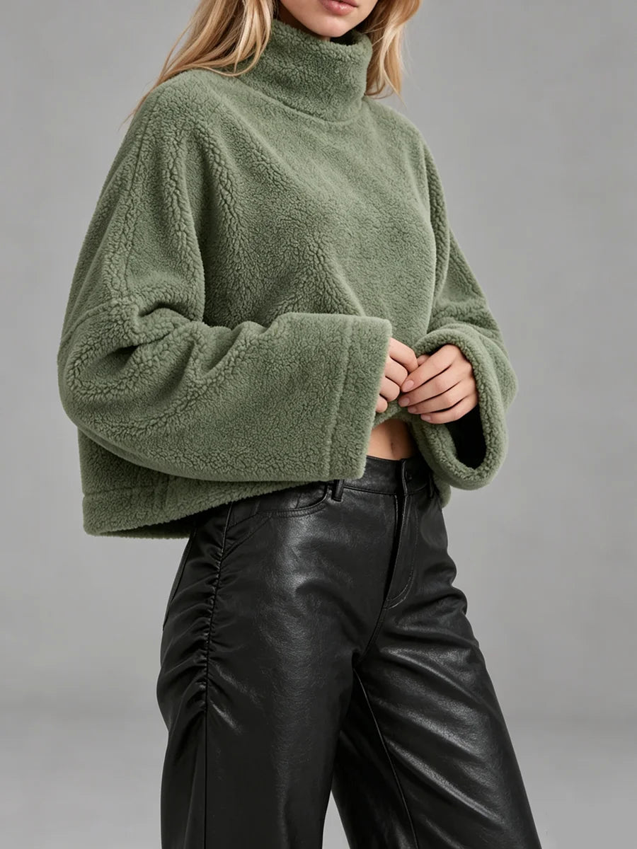 A woman wearing a cropped, oversized green sherpa fleece turtleneck sweater and black faux-leather pants, photographed against a plain gray studio background.
