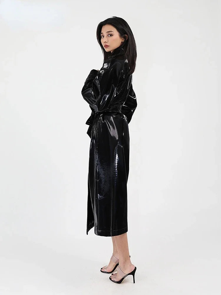 A woman modeling a glossy black long trench coat with a matching waist tie, paired with black high-heeled sandals, against a plain white studio background.