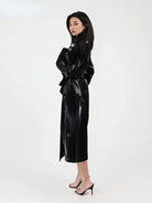 A woman modeling a glossy black long trench coat with a matching waist tie, paired with black high-heeled sandals, against a plain white studio background.