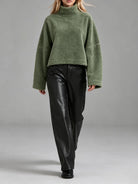 A model wearing an oversized, cropped green fleece turtleneck sweater and black leather pants, walking against a neutral gray studio background.