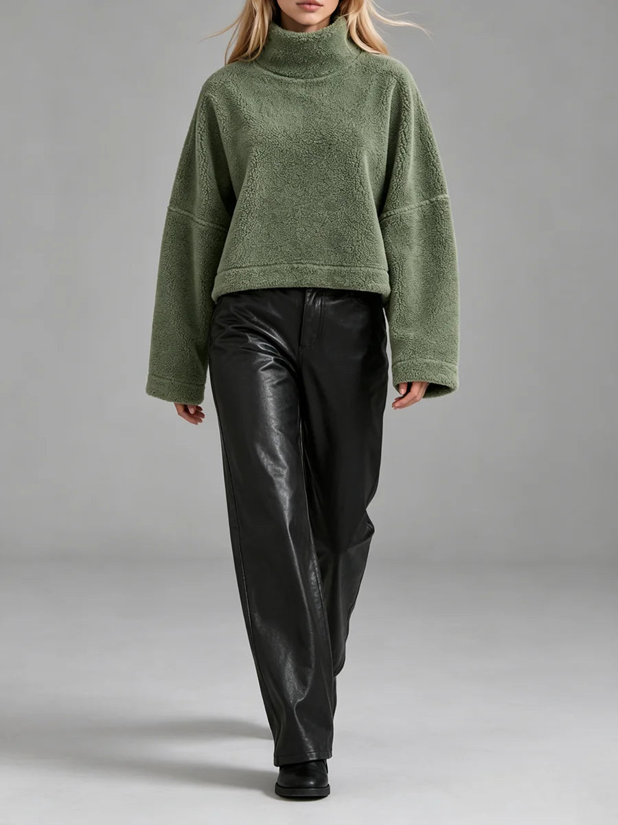 A model wearing an oversized, cropped green fleece turtleneck sweater and black leather pants, walking against a neutral gray studio background.