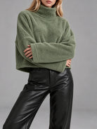 A woman wearing an oversized sage green fleece turtleneck sweater and black leather pants, shown from the neck down against a plain gray studio background.