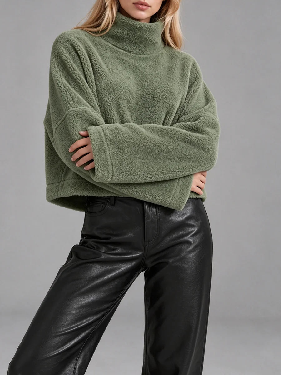 A woman wearing an oversized sage green fleece turtleneck sweater and black leather pants, shown from the neck down against a plain gray studio background.